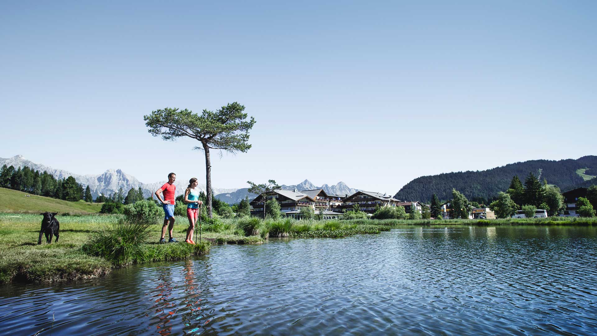 Paar mit Stöcken am Ufer des Sees vor Hotel und Wald unter klarem Himmel.