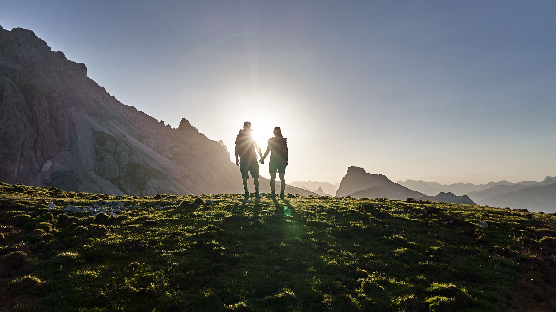Paar hält Händchen auf Almwiese im Sonnenaufgang vor majestätischer Bergkulisse.