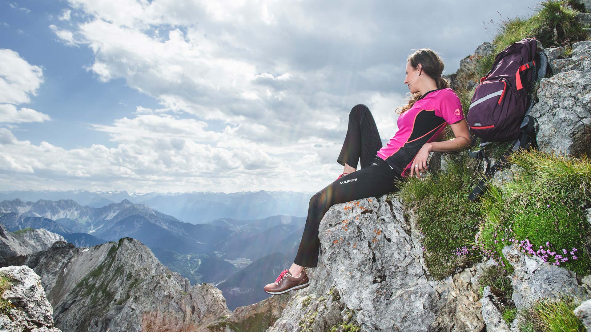 Frau mit Rucksack ruht auf felsigem Grat und blickt auf weite Berglandschaft unter Wolken.